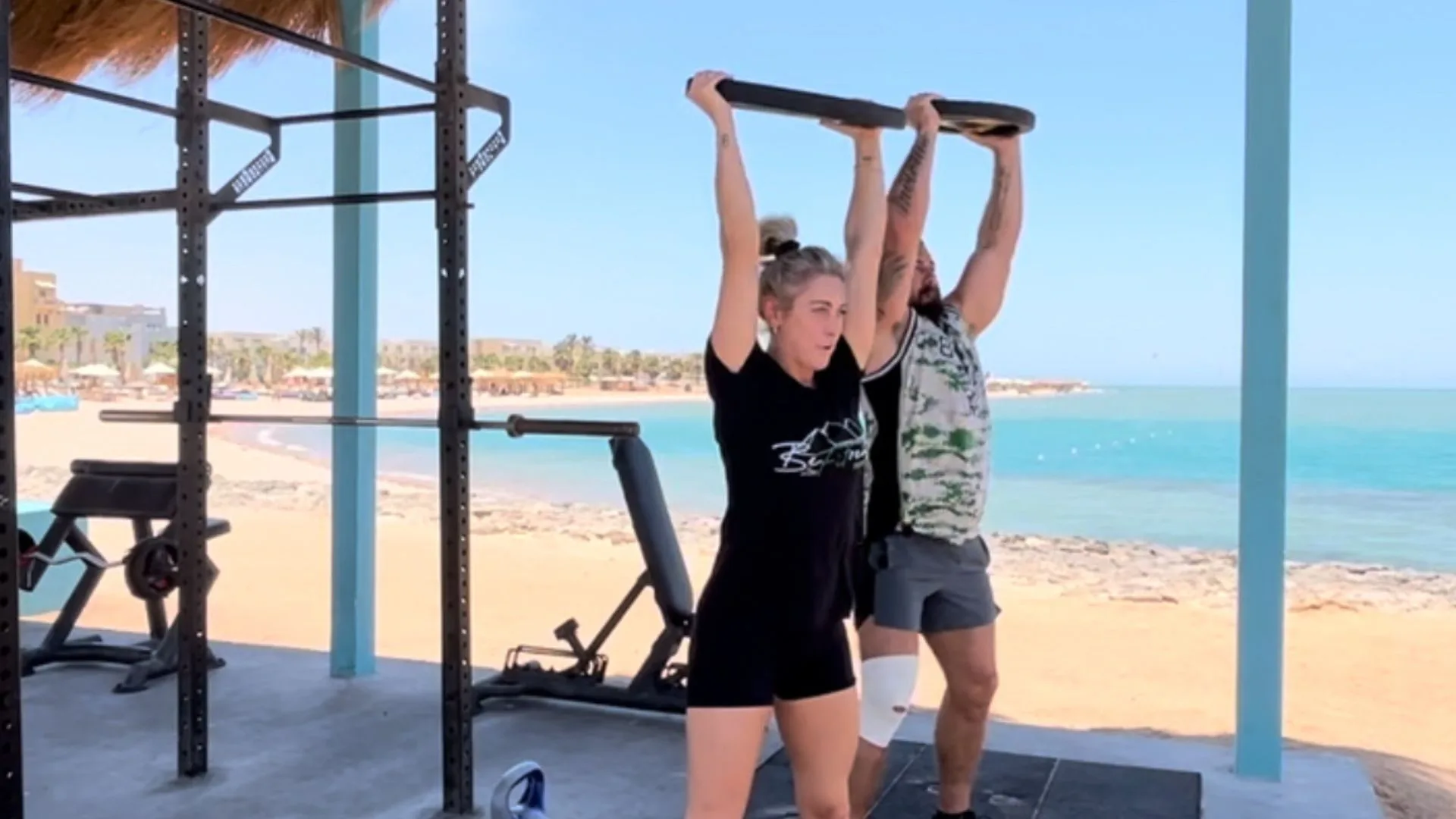 A male and female couple at an outdoor beach gym lifting a weight plate over their heads, embodying the spirit of a fitness retreat for couples with teamwork and an active lifestyle in a stunning coastal setting.