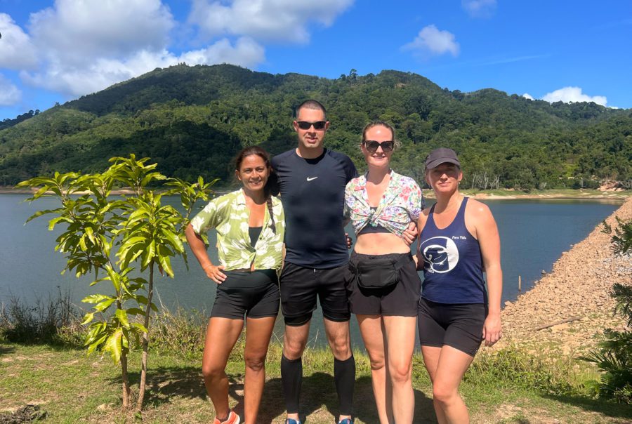 a group of three females and one male standing together with their arms around each other and the sea and mountains in the background