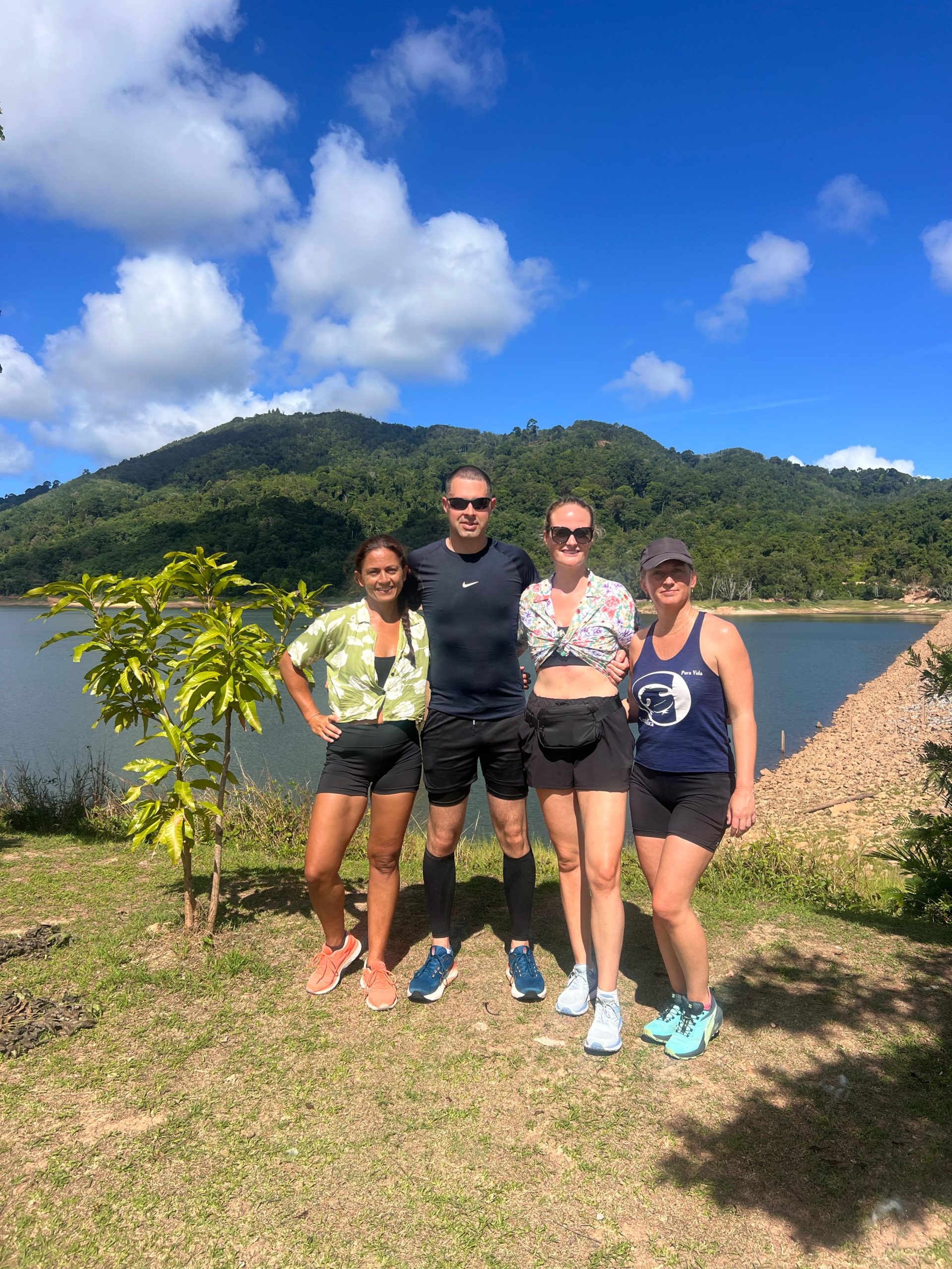 a group of three females and one male standing together with their arms around each other and the sea and mountains in the background